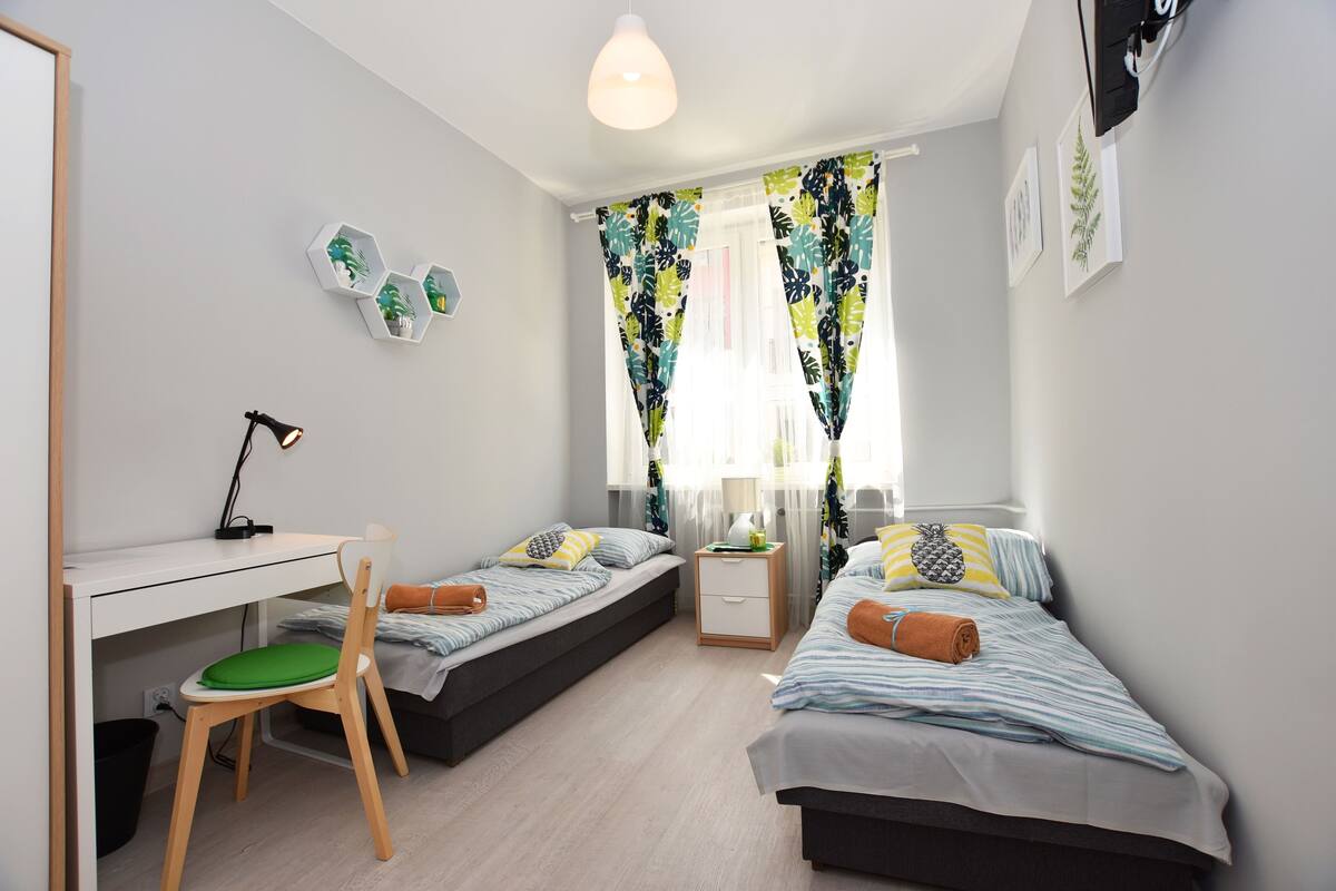 A bedroom with two single beds, a desk with a chair, a nightstand, and a window with curtains.