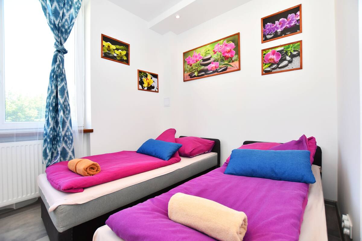A hotel room with two beds, each with a different color bedspread and pillows. There are wall-mounted pictures of flowers and a window with a curtain.