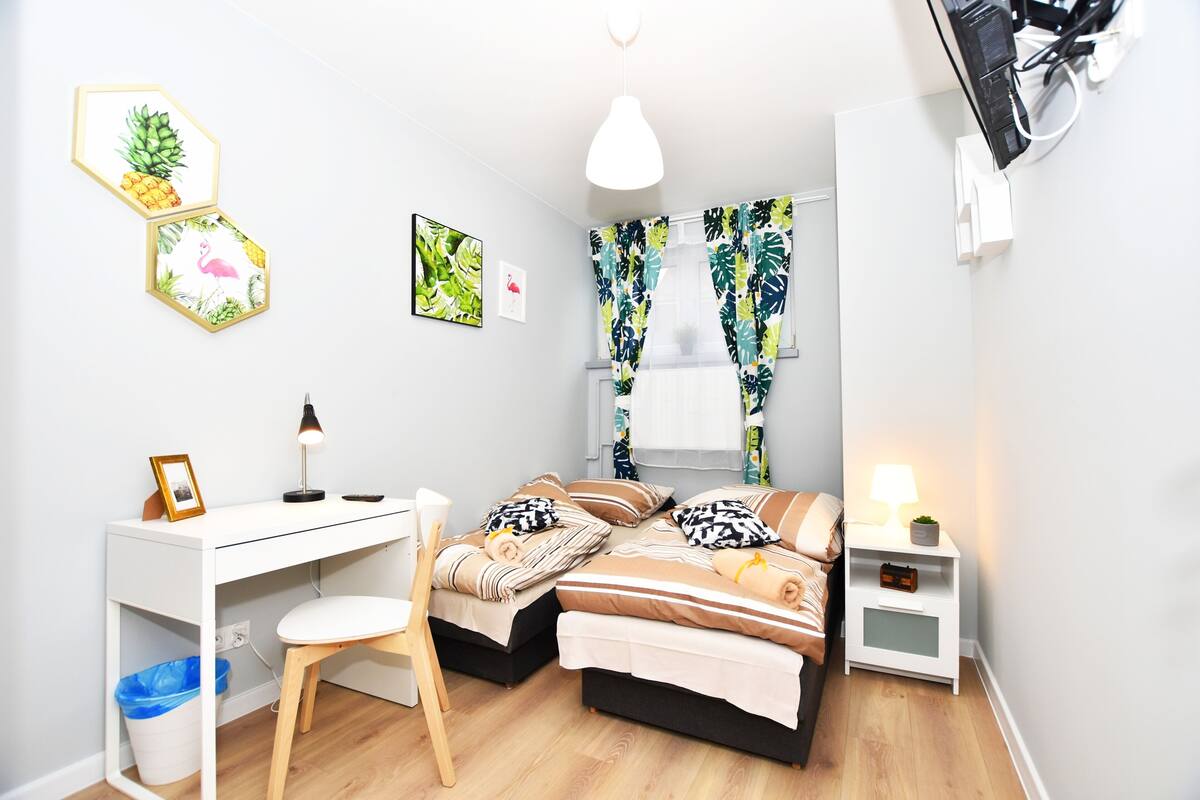 A modern bedroom with a desk, two beds, a chair, and a nightstand.