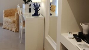 Ett kompakt hotellrum med ett vitt skrivbord, en stol, en liten orkidé i en kruka, en lampa, en kaffebryggare, en fjärrkontroll och en inramad tavla på väggen.