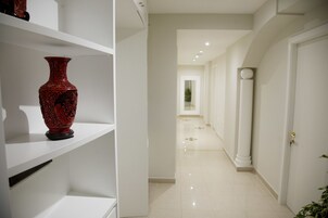 Um corredor com uma porta branca, uma estante branca com um vaso vermelho e piso de azulejos.
