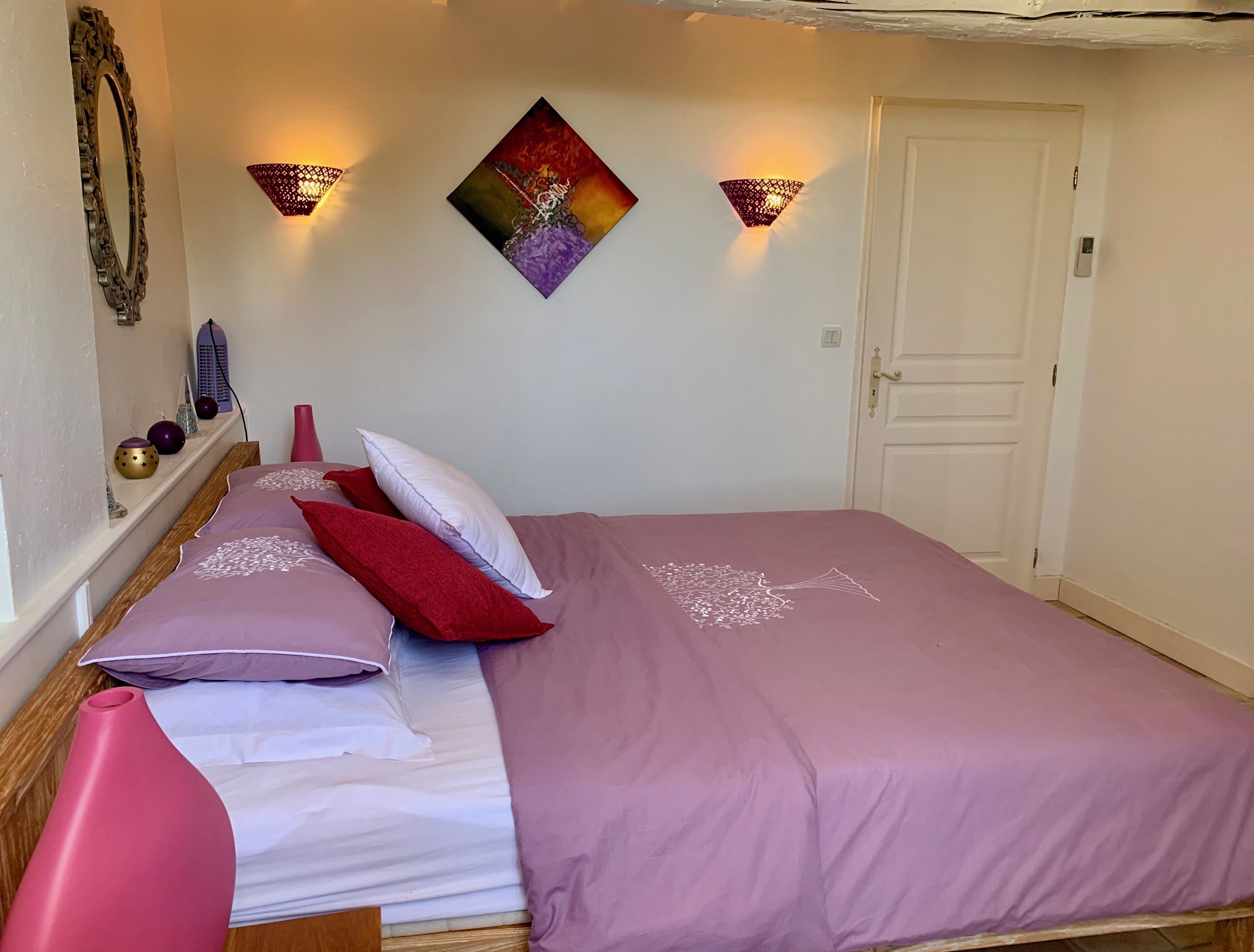 A neatly made bed with a purple bedspread and white linens, a pink vase, and a decorative wall piece.