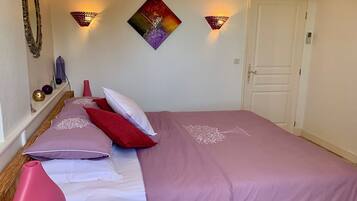A neatly made bed with a purple bedspread and white linens, a pink vase, and a decorative wall piece.