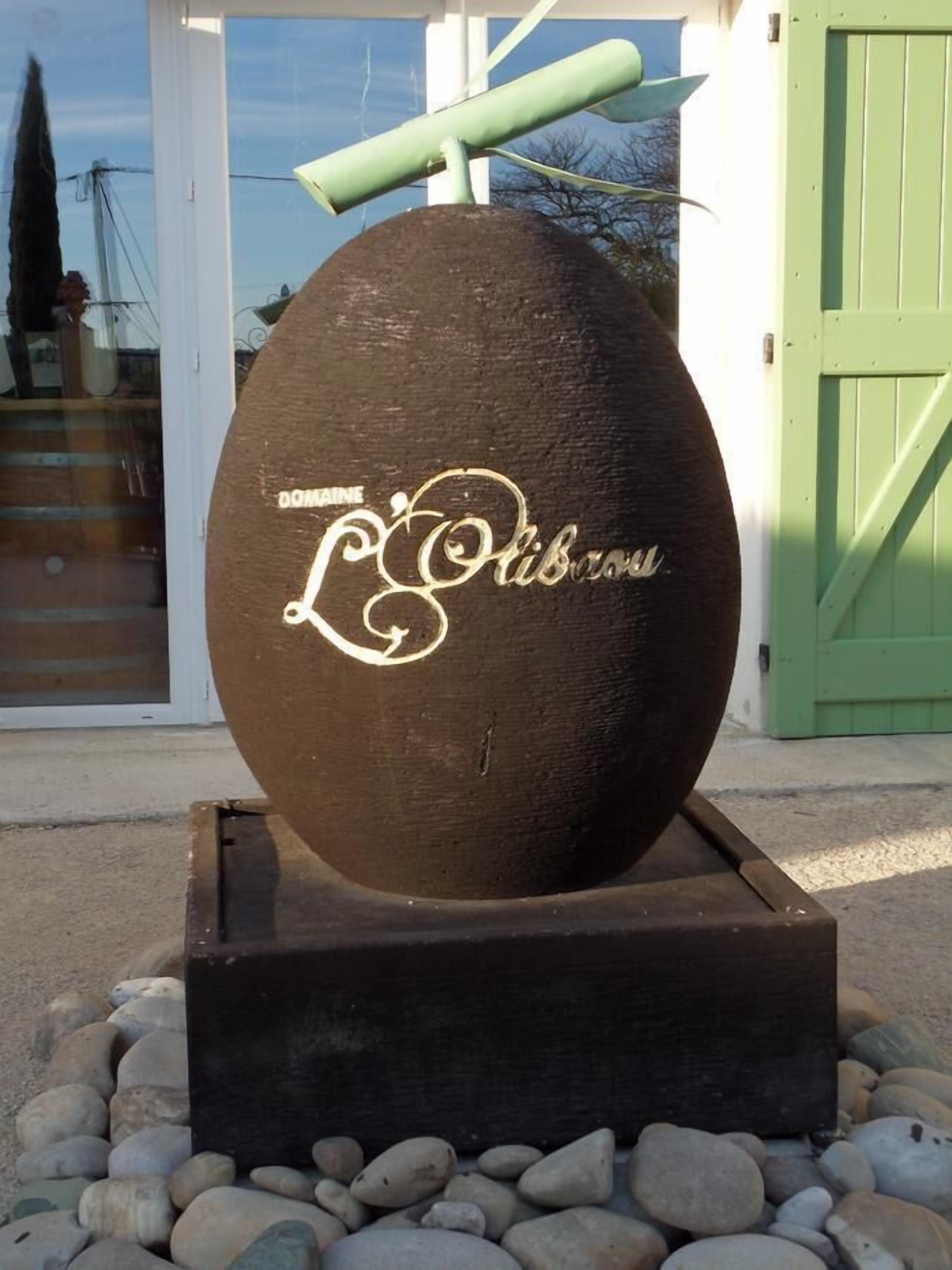 A large, spherical object with the inscription "DOMAINE Libboux" on a brown surface, placed on a black base surrounded by rocks.