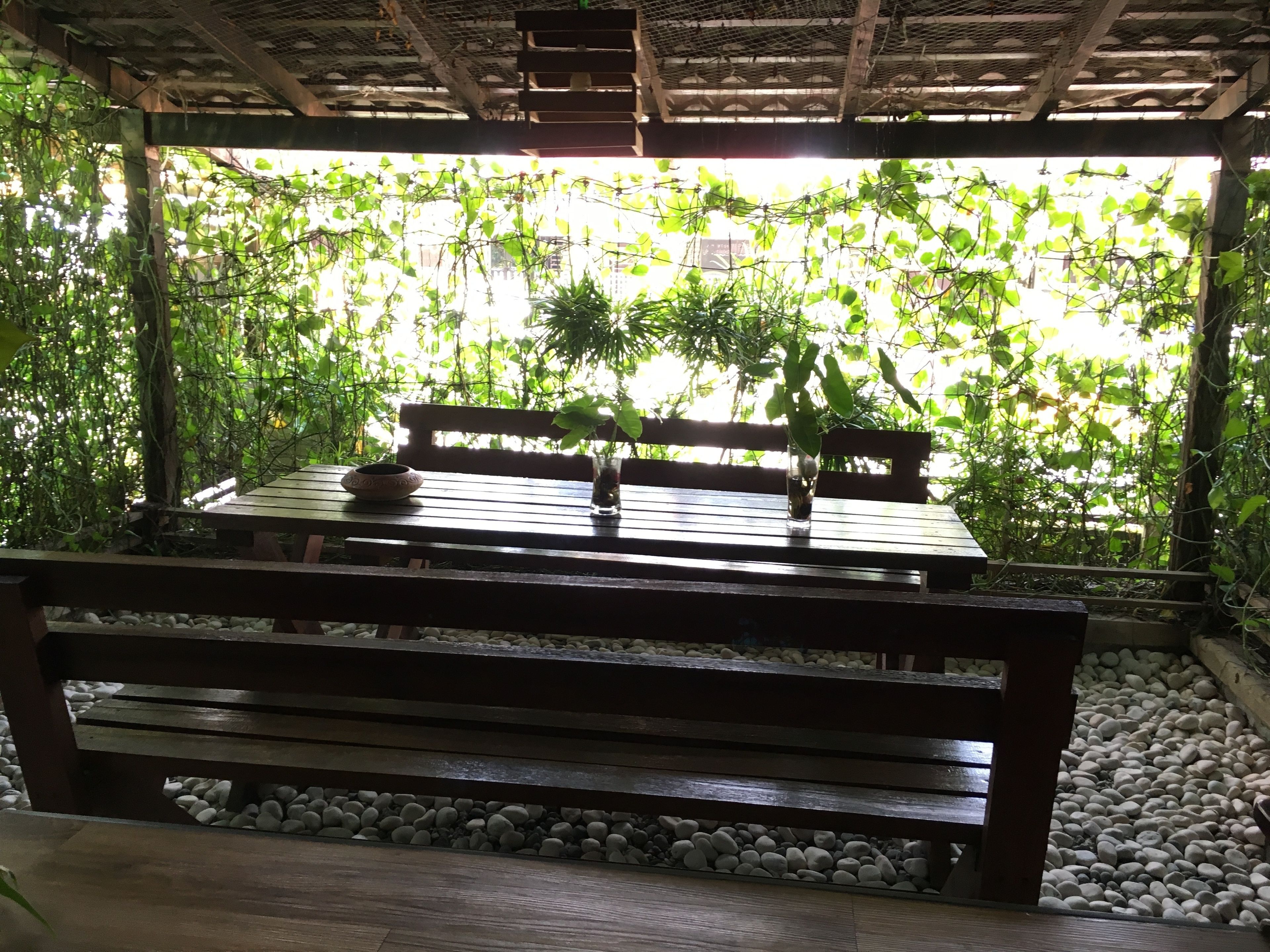 Ein Holztisch mit einer Schale und einer Glasvase, platziert auf einer Bank unter einer Pergola mit Blick auf dichtes Grün.
