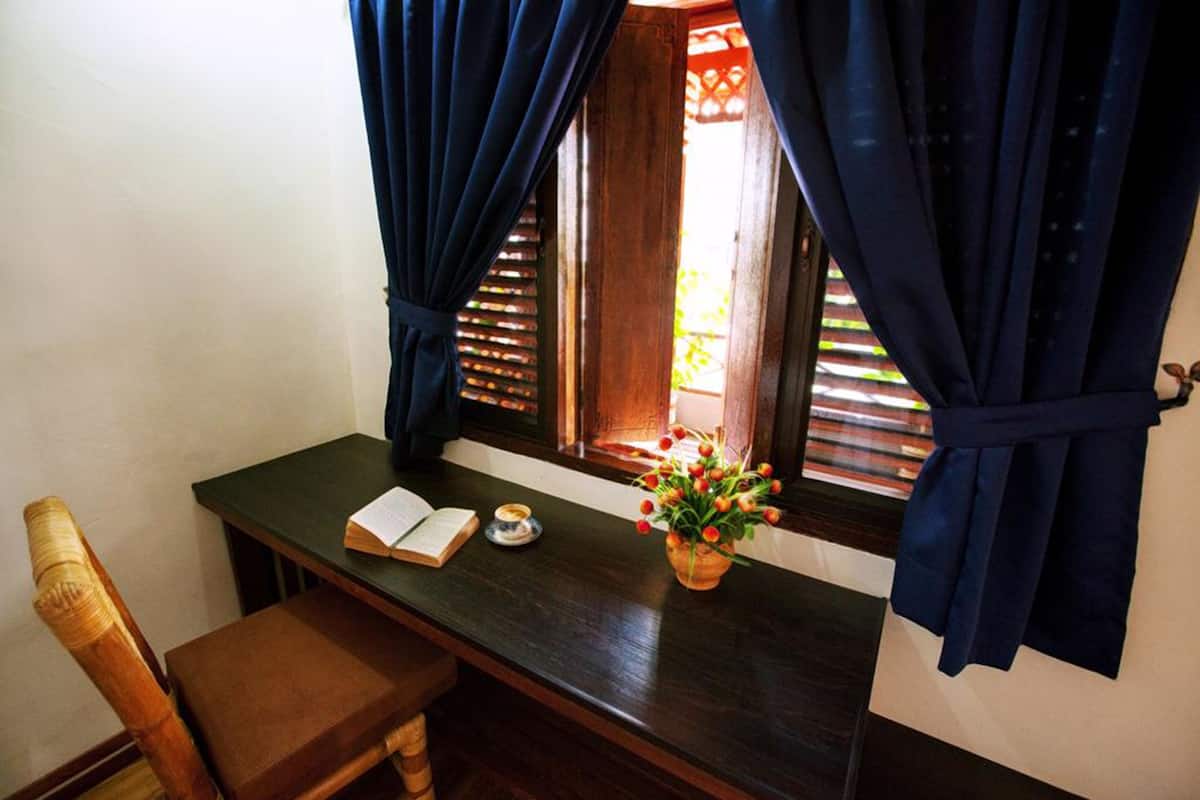 A wooden desk with a book, a small bowl, and a vase with flowers by a window with dark curtains.