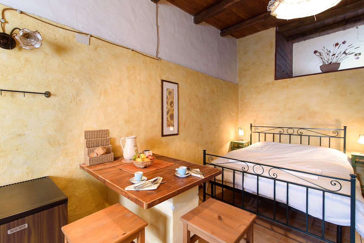 Un dormitorio con estructura de cama de madera, una mesita de noche con lámpara, y una pequeña mesa con una cesta de fruta y una jofaina.