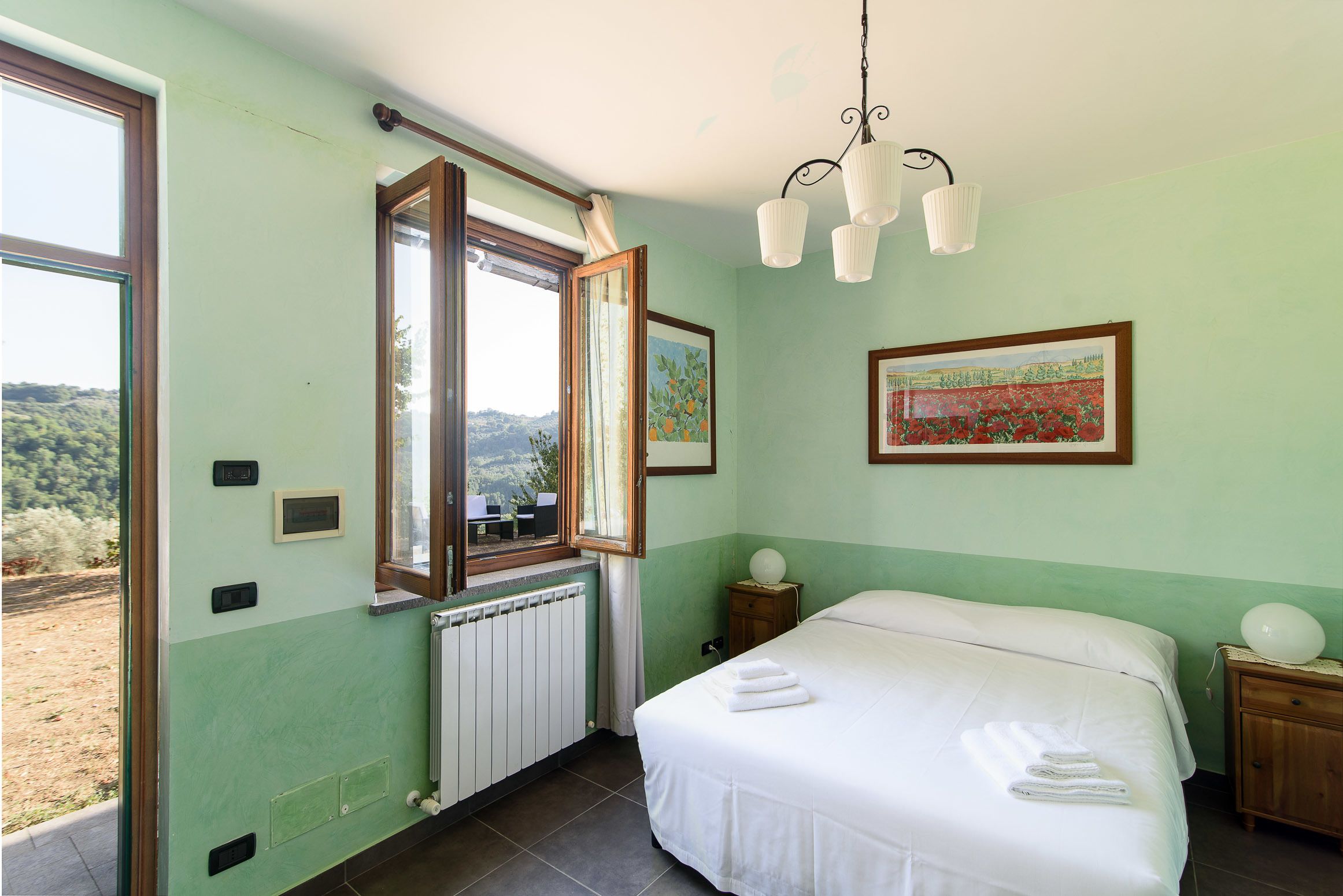 Een slaapkamer met een groot bed, een raam met een open luik, een radiator en een wandlamp.