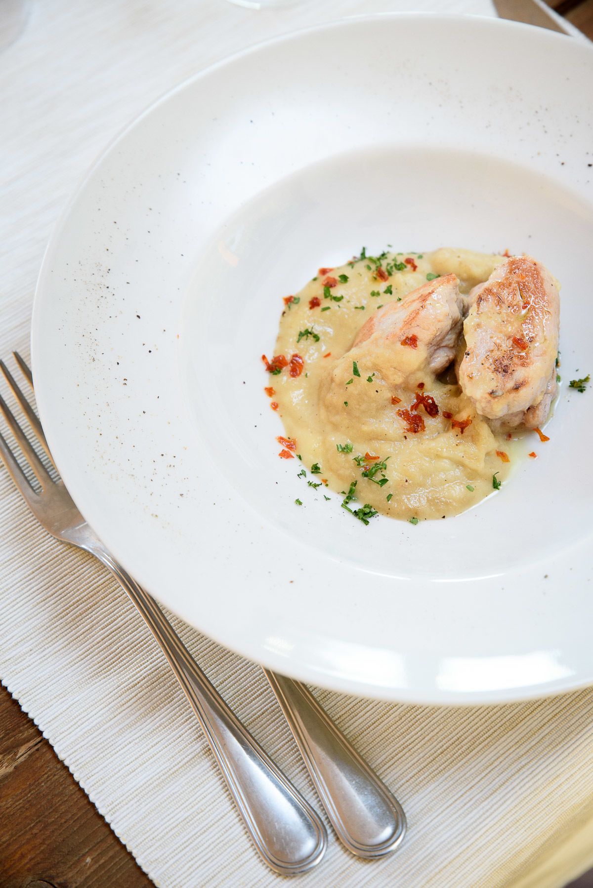 A white plate with a piece of chicken and a creamy sauce, garnished with herbs, on a wooden table with a white cloth.