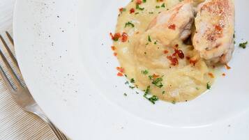 Ein weißer Teller mit Hähnchenstück und cremiger Soße, garniert mit Kräutern, auf einem Holztisch mit weißer Tischdecke.