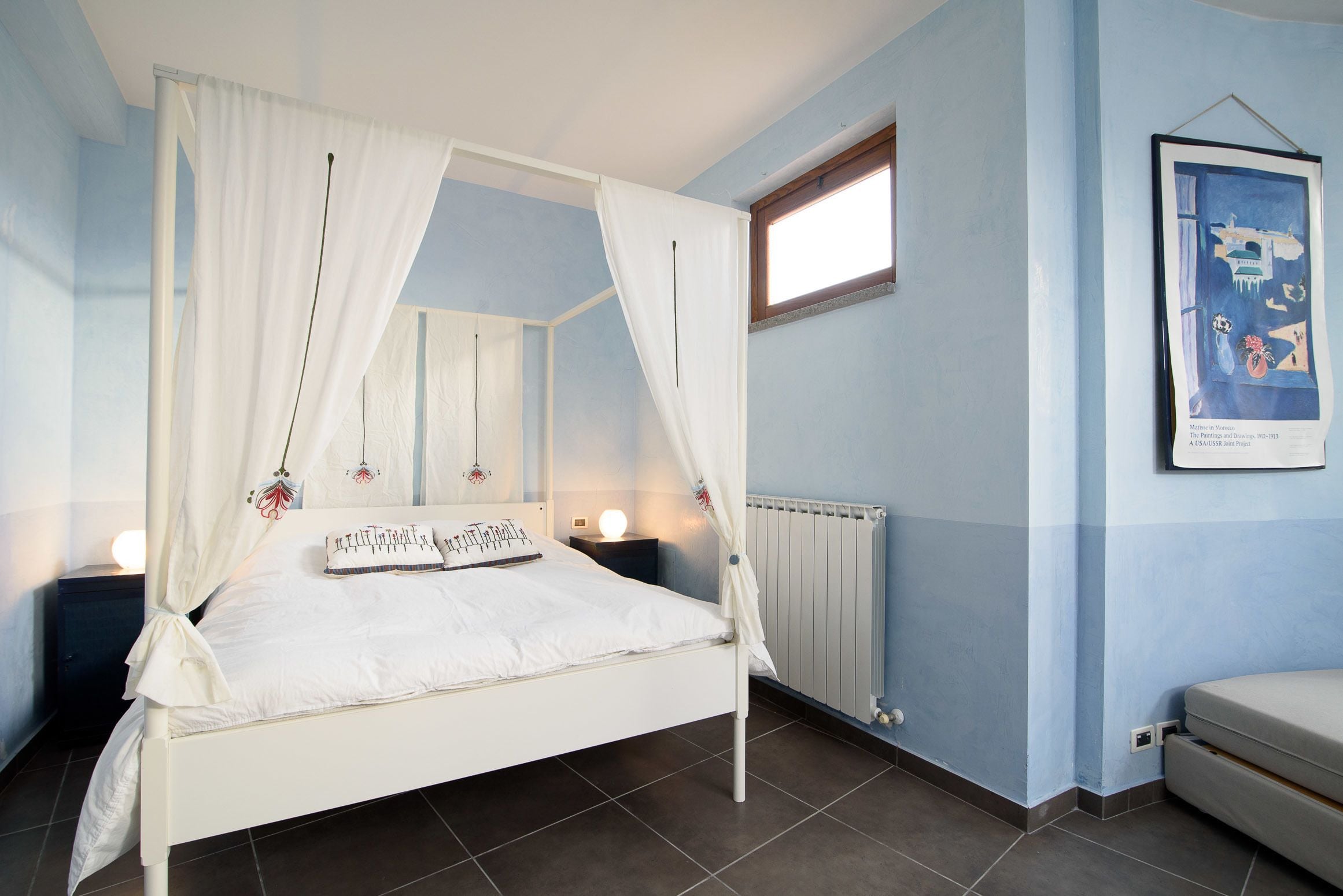 A bedroom with a four-poster bed, a nightstand, a bedside table, a framed picture on the wall, and a window.