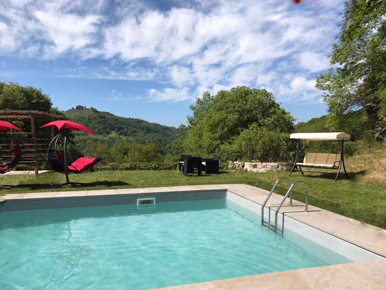 Ein rechteckiger Swimmingpool mit klarem, blauem Wasser, umgeben von einer Betonplattform. In der Nähe des Pools befinden sich zwei rote Sonnenschirme und eine schwarze Liege.