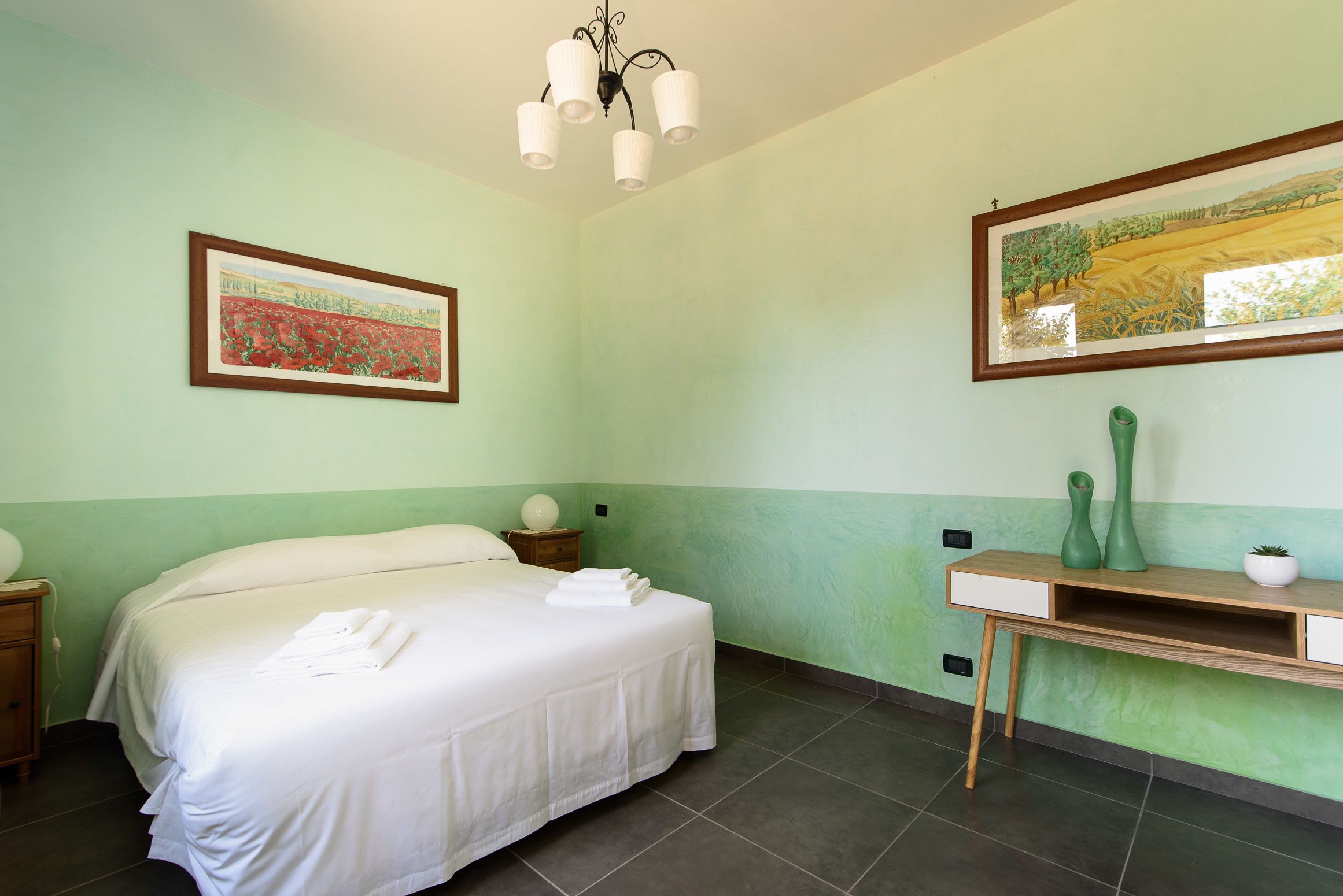 A neatly made bed with white linens and towels, a wooden nightstand with a lamp, a desk with a vase and a plant, and two framed artworks on the walls.