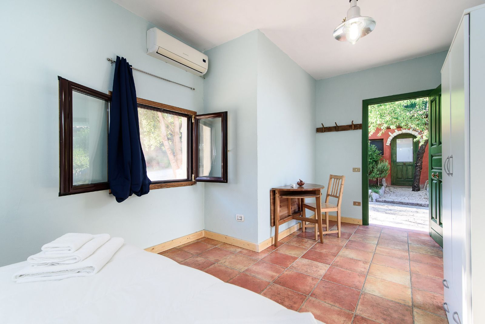 A room with a bed, a table with a chair, an air conditioning unit, and a view of a green door leading outside.