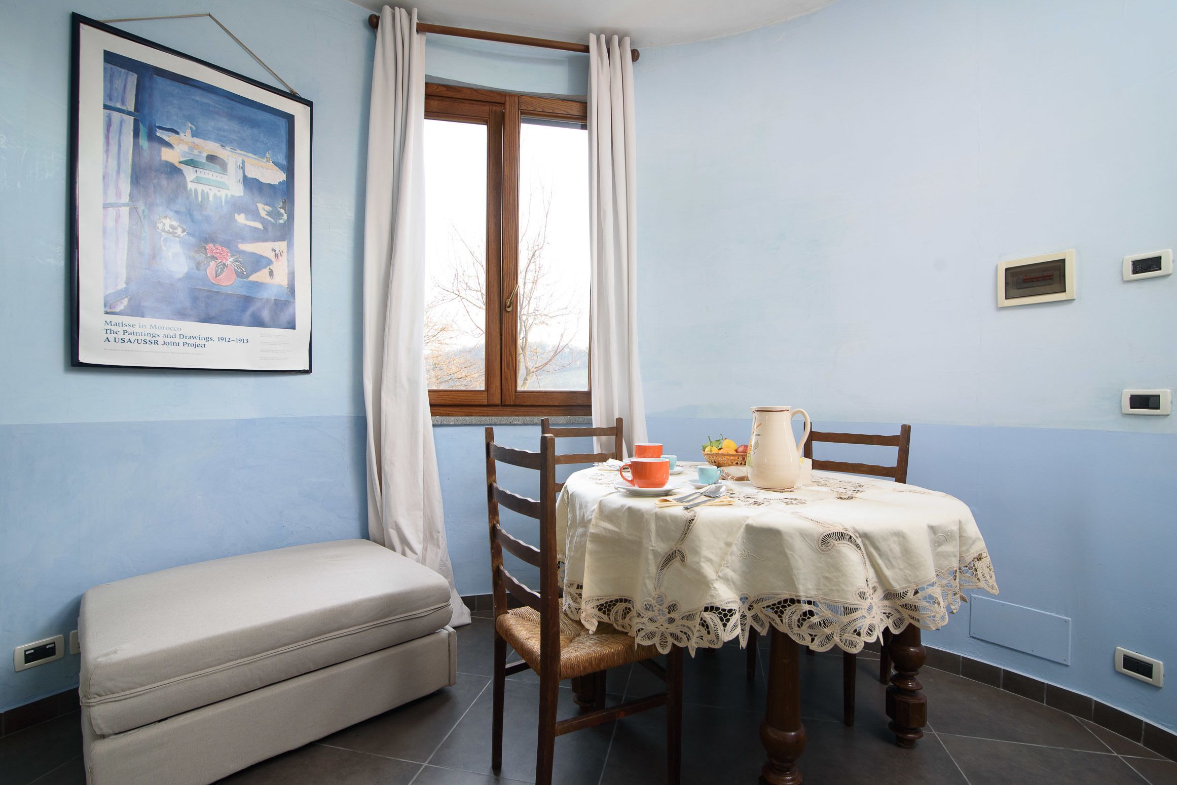 A room with a dining table set for two, a bench, a window with curtains, and a painting on the wall.