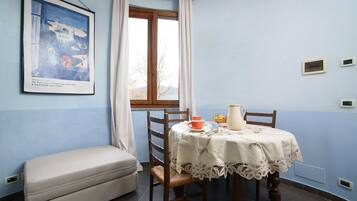 A room with a dining table set for two, a bench, a window with curtains, and a painting on the wall.