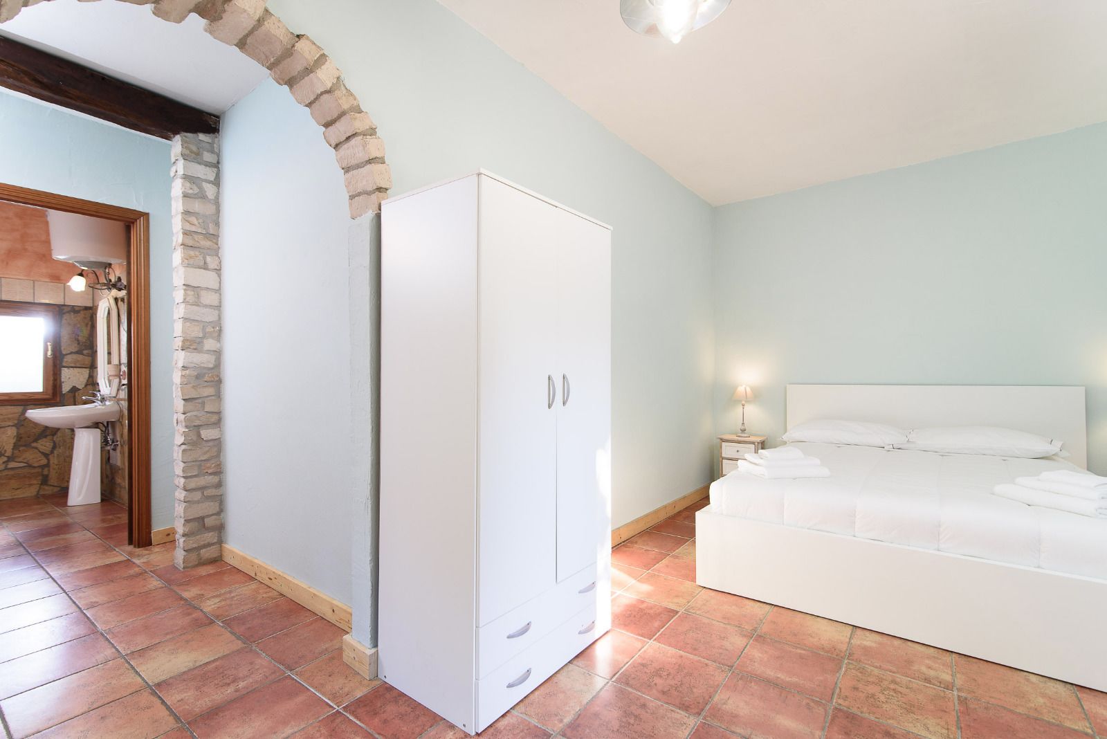 A bedroom with a bed, a wardrobe, a bathroom with a sink, and a stone archway.