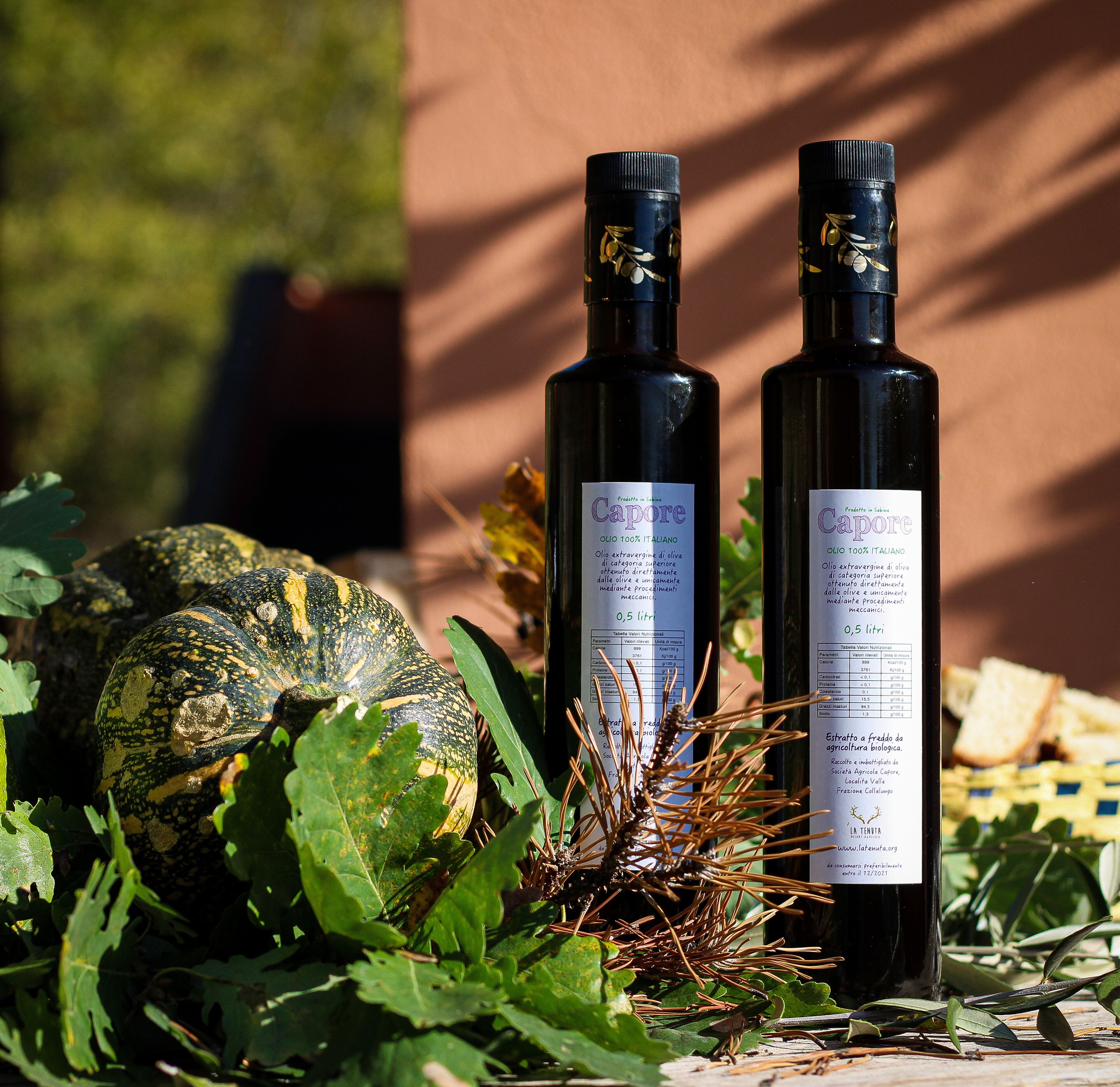 Two bottles of Capore olive oil, a large pumpkin, and pine cones on a table with greenery.