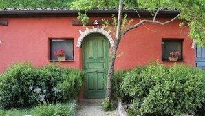 Un edificio rojo con una puerta verde, dos ventanas con macetas de flores y un camino de grava que lleva a la puerta.