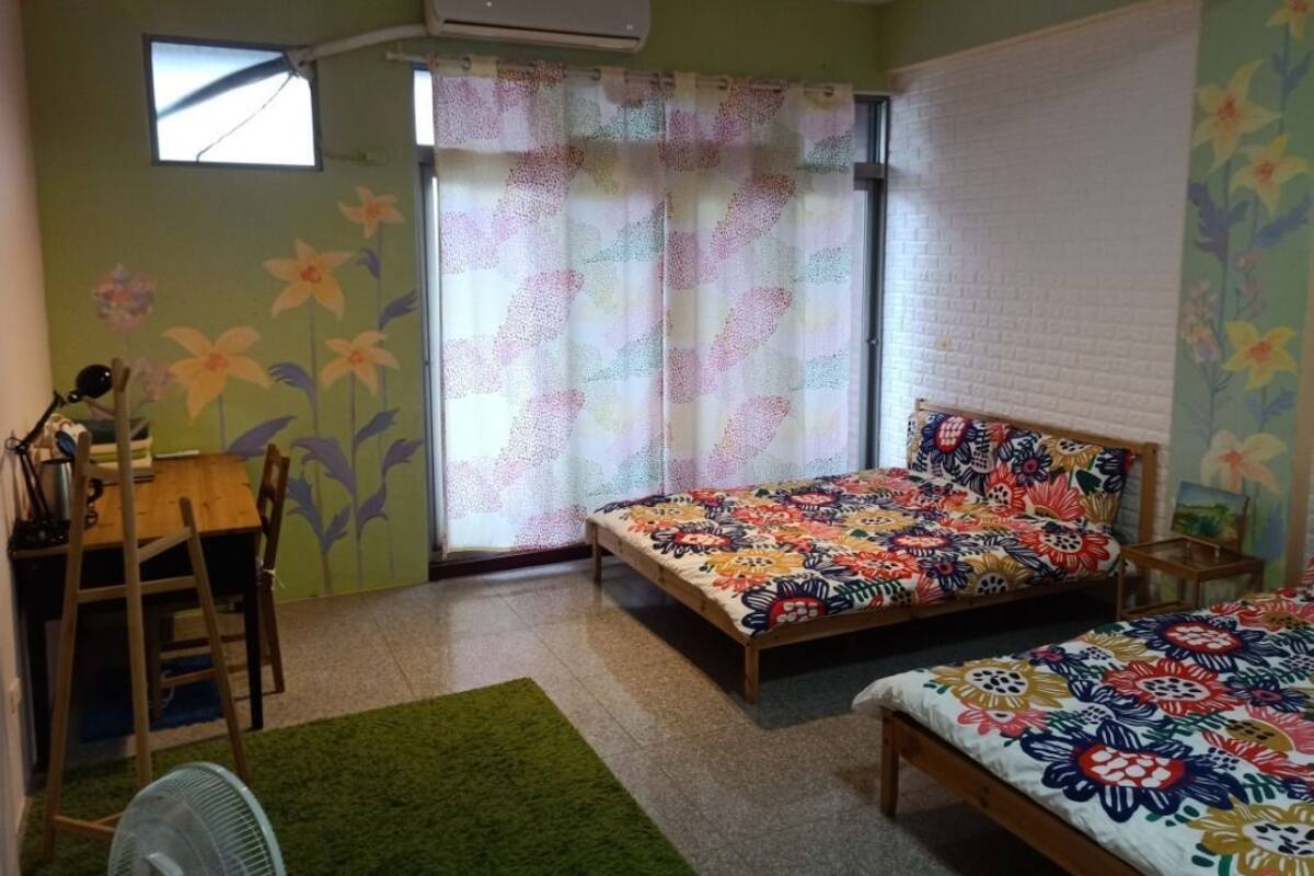A bedroom with a bed, a desk, and a chair. There is a window with a curtain, an air conditioner, and a green rug.
