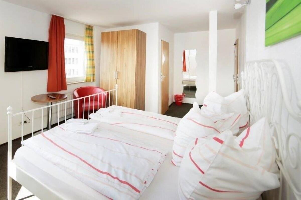 Hotellihuone, jossa on sänky, televisio, punainen tuoli ja puinen vaatekaappi.