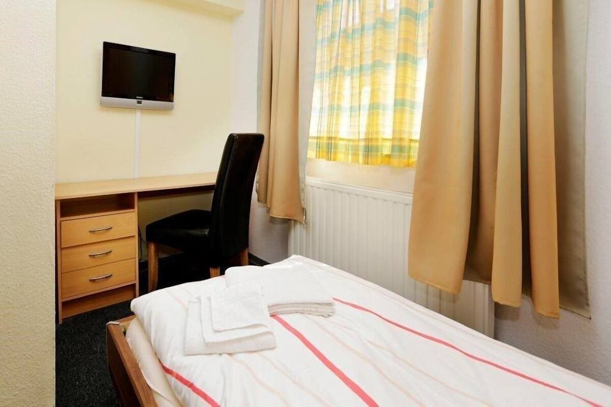 Hotellihuone, jossa on sänky, työpöytä tuolilla, seinälle asennettu televisio ja ikkuna, jossa on verhot.