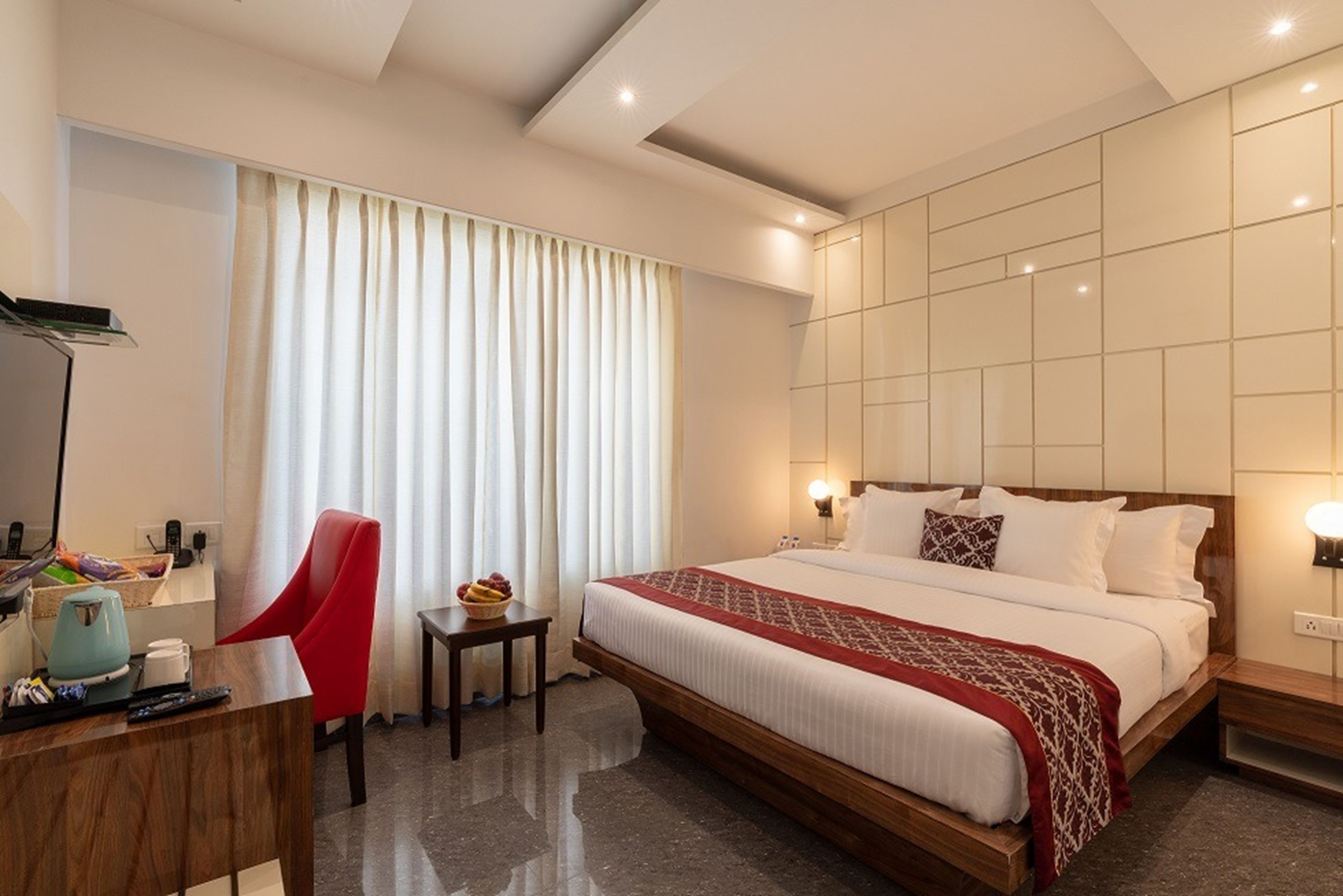 A hotel room with a large bed, a desk with a chair, a small table with a fruit bowl, and a large window with curtains.