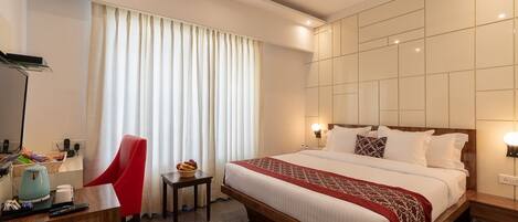 A hotel room with a large bed, a desk with a chair, a small table with a fruit bowl, and a large window with curtains.