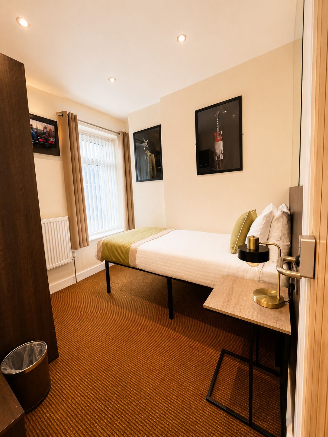 A hotel room with a bed, a bedside table with a lamp, a window with curtains, a radiator, and a TV mounted on the wall.