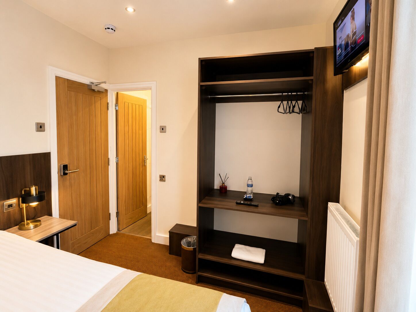 A hotel room with a bed, a wooden wardrobe, a small table with a lamp, a television, and a radiator.