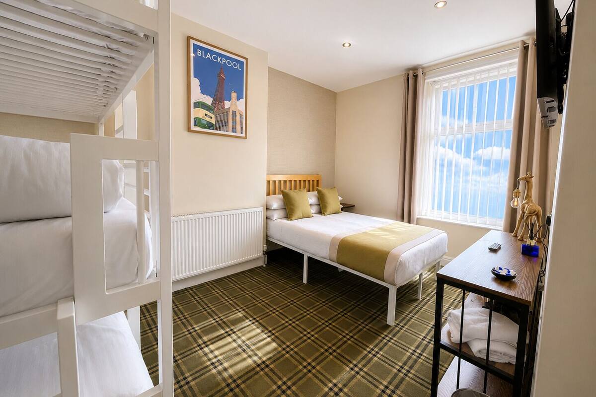 A hotel room with a bed, a desk, a radiator, a window with curtains, a poster on the wall, and a television mounted on the wall.