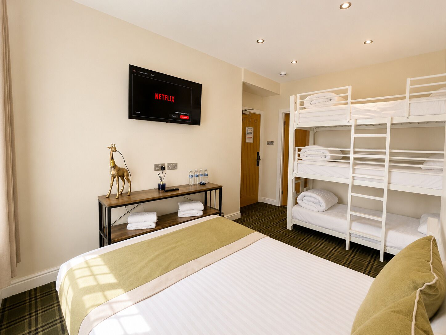 A hotel room with a bed, a TV mounted on the wall, a bedside table with a lamp, a bench with towels, and a bunk bed.
