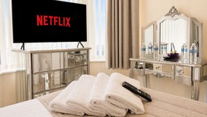Quarto de hotel com cama, televisão exibindo 'NETFLIX', penteadeira com espelho e janela com cortinas.