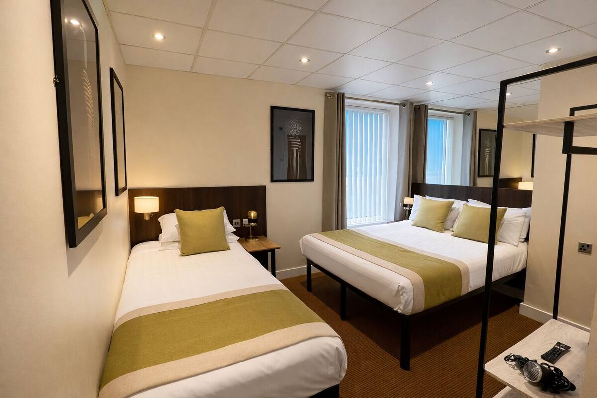 A hotel room with two beds, a bedside table, a lamp, a mirror, and a window with curtains.