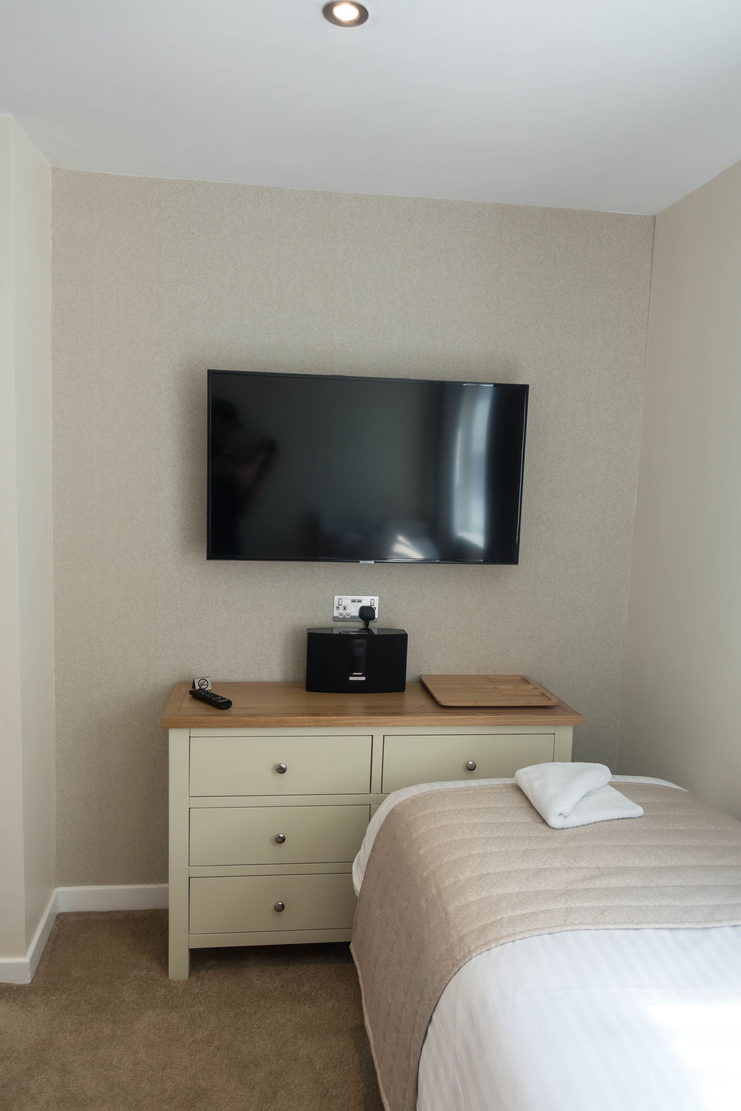 A hotel room with a flat-screen TV mounted on the wall, a wooden dresser with drawers, a remote control, and a neatly made bed with a white comforter.