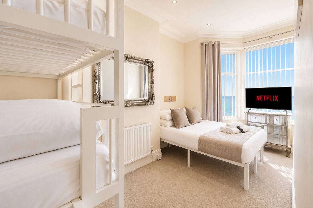 A hotel room with bunk beds, a bed with a TV displaying 'NETFLIX,' a window with curtains, and a radiator.