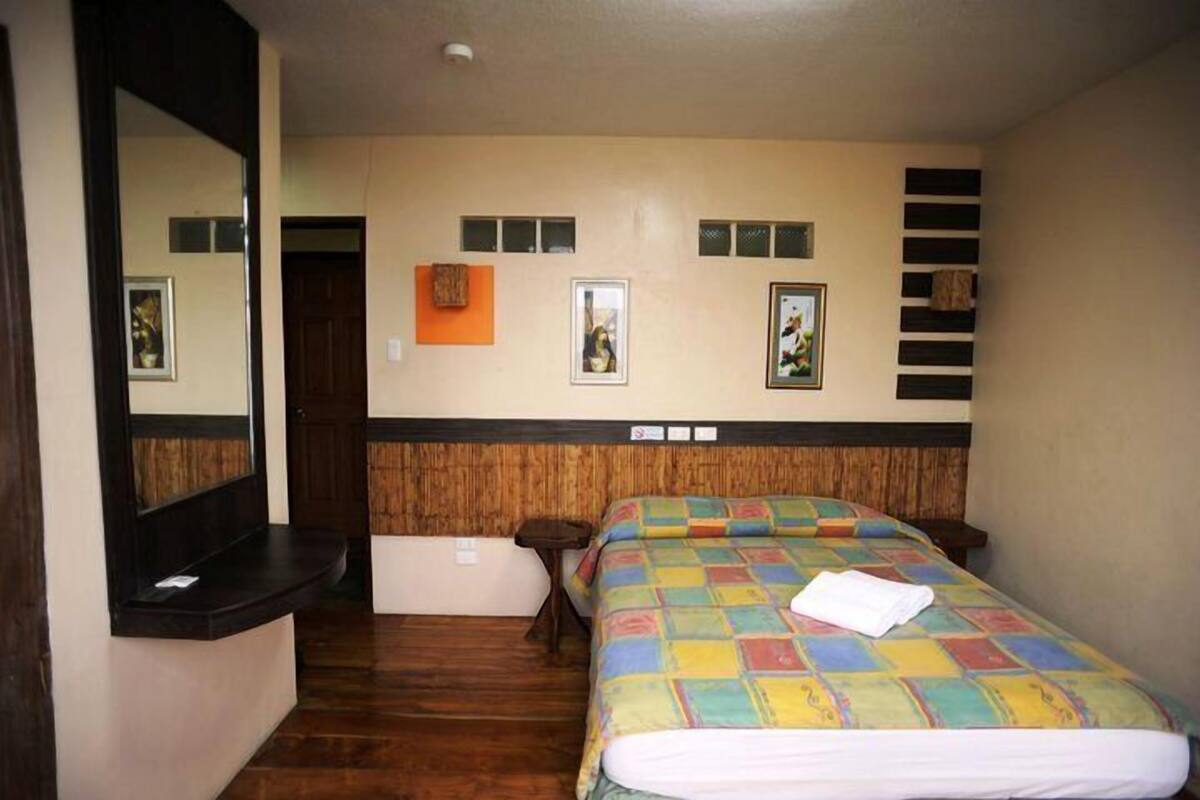 A hotel room with a wooden floor, a bed with a colorful quilt, a small bedside table, a mirror, and a door.