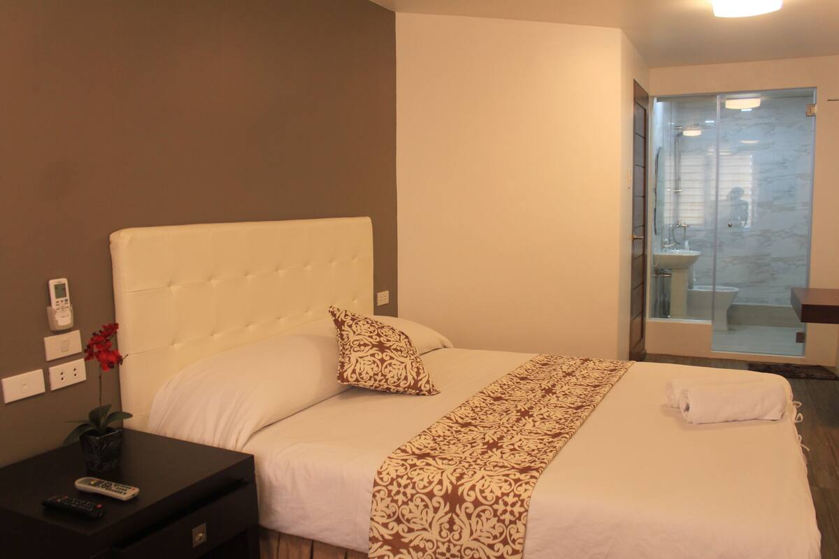 A hotel room with a large bed, a bedside table with a remote control, a vase with flowers, and a bathroom with a glass door.