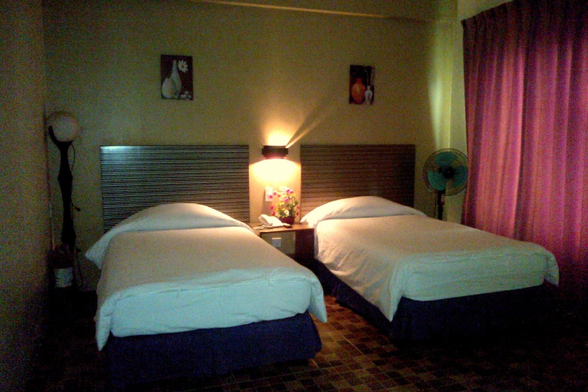 A hotel room with two beds, a bedside table with a lamp, a fan, and a wall-mounted picture.