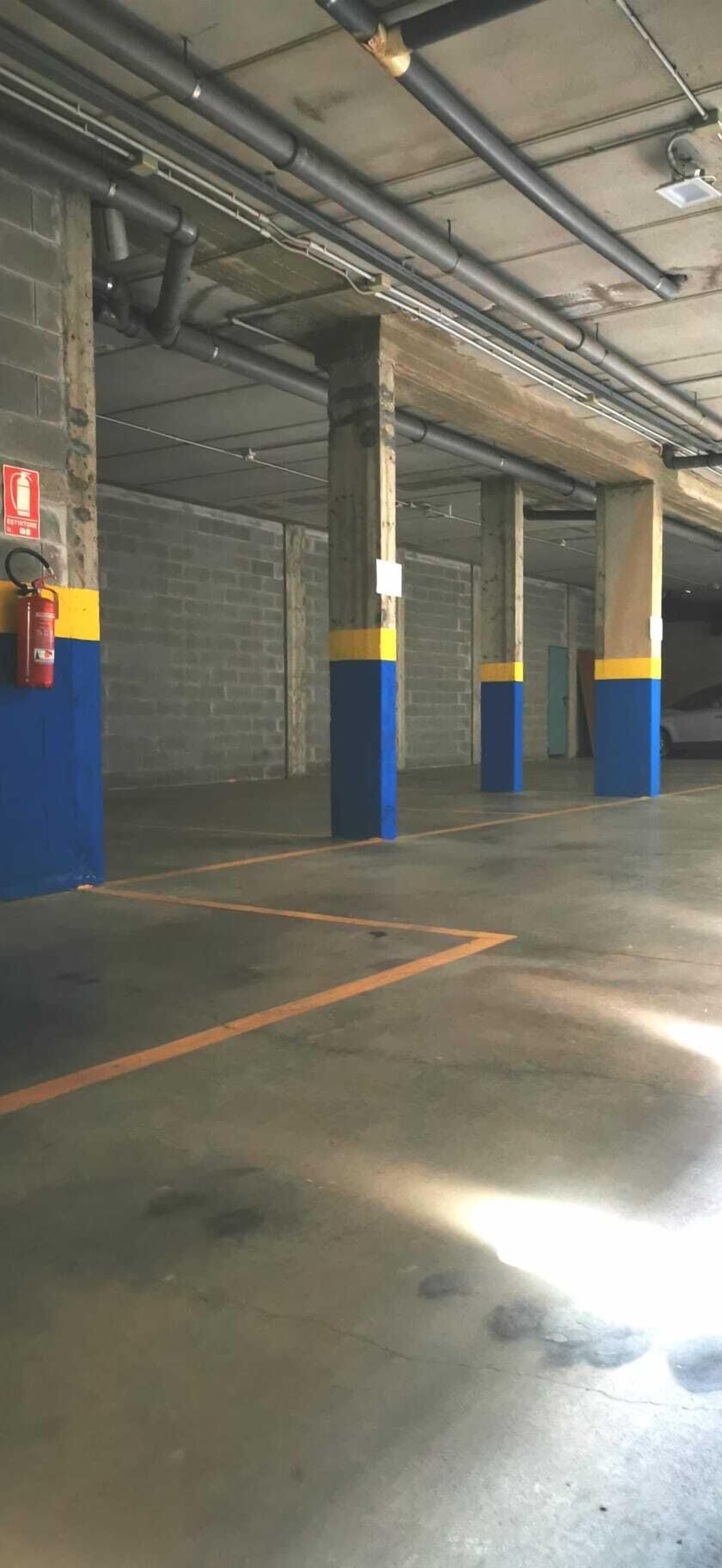 Un parking souterrain avec des piliers en béton, des lignes de stationnement orange et un extincteur fixé au mur.