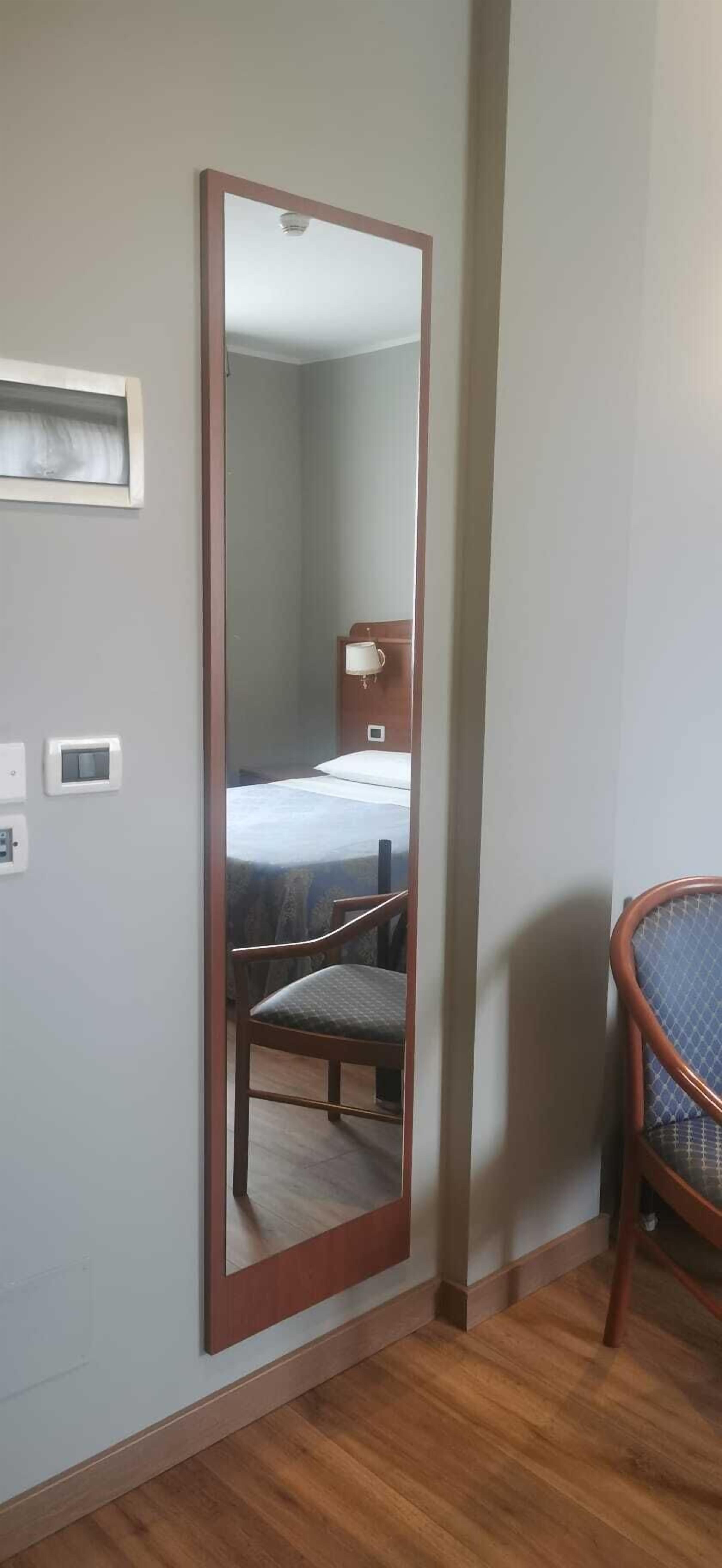 A room with a large mirror, a bed with a white cover, a chair, and a bedside table with a lamp.