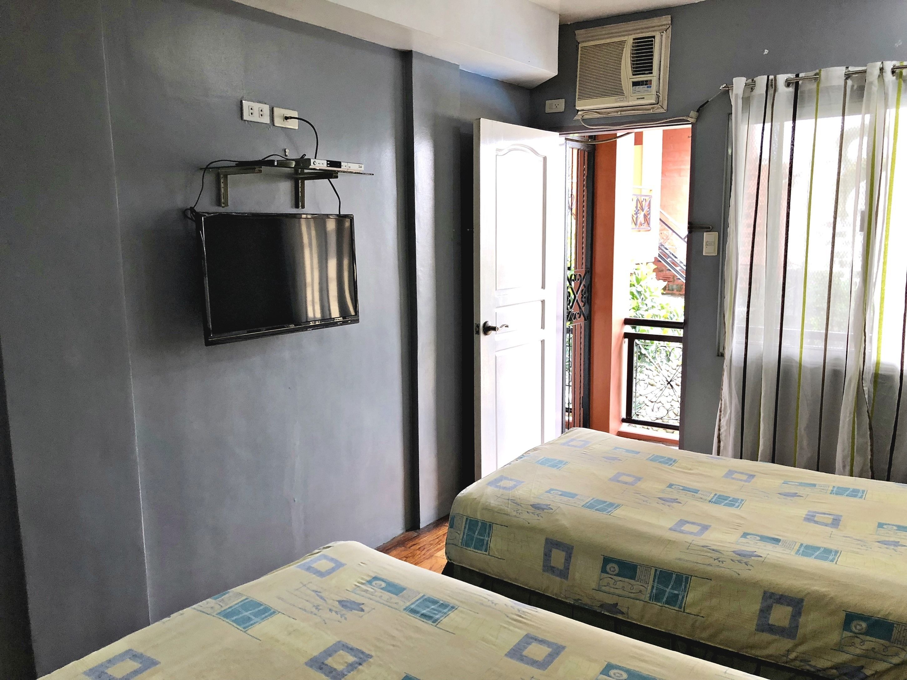 A room with two beds, a flat-screen TV mounted on the wall, an air conditioning unit, and a door leading to an outdoor balcony.