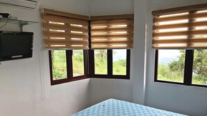 A bedroom with a bed, two windows with blinds, a TV on the wall, and a view of greenery outside. - Pura Vida Villas (Tagaytay)