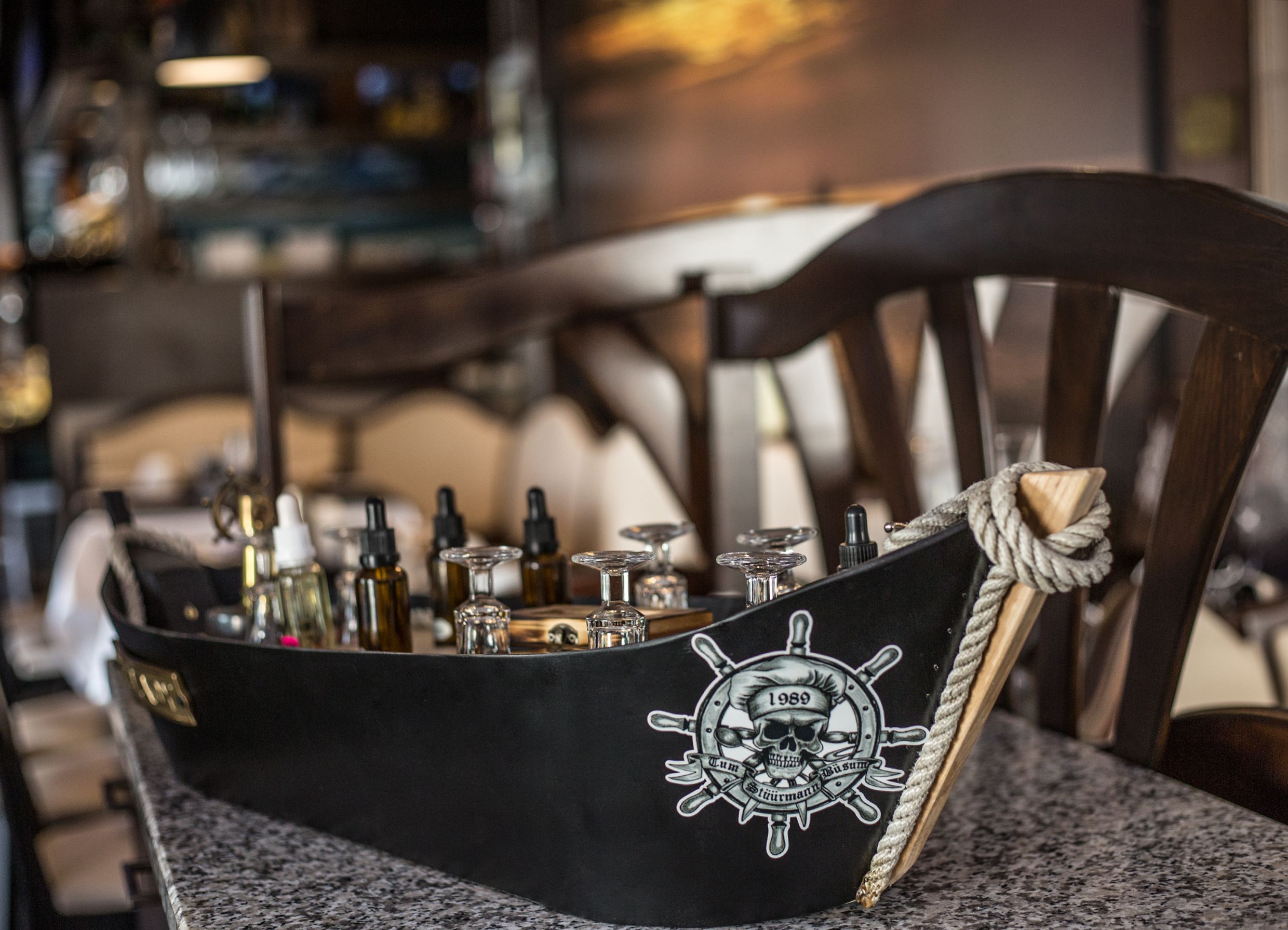 A ship-shaped bar with bottles and glasses, a rope tied to the backrest of a chair, and a skull emblem with the year 1989.
