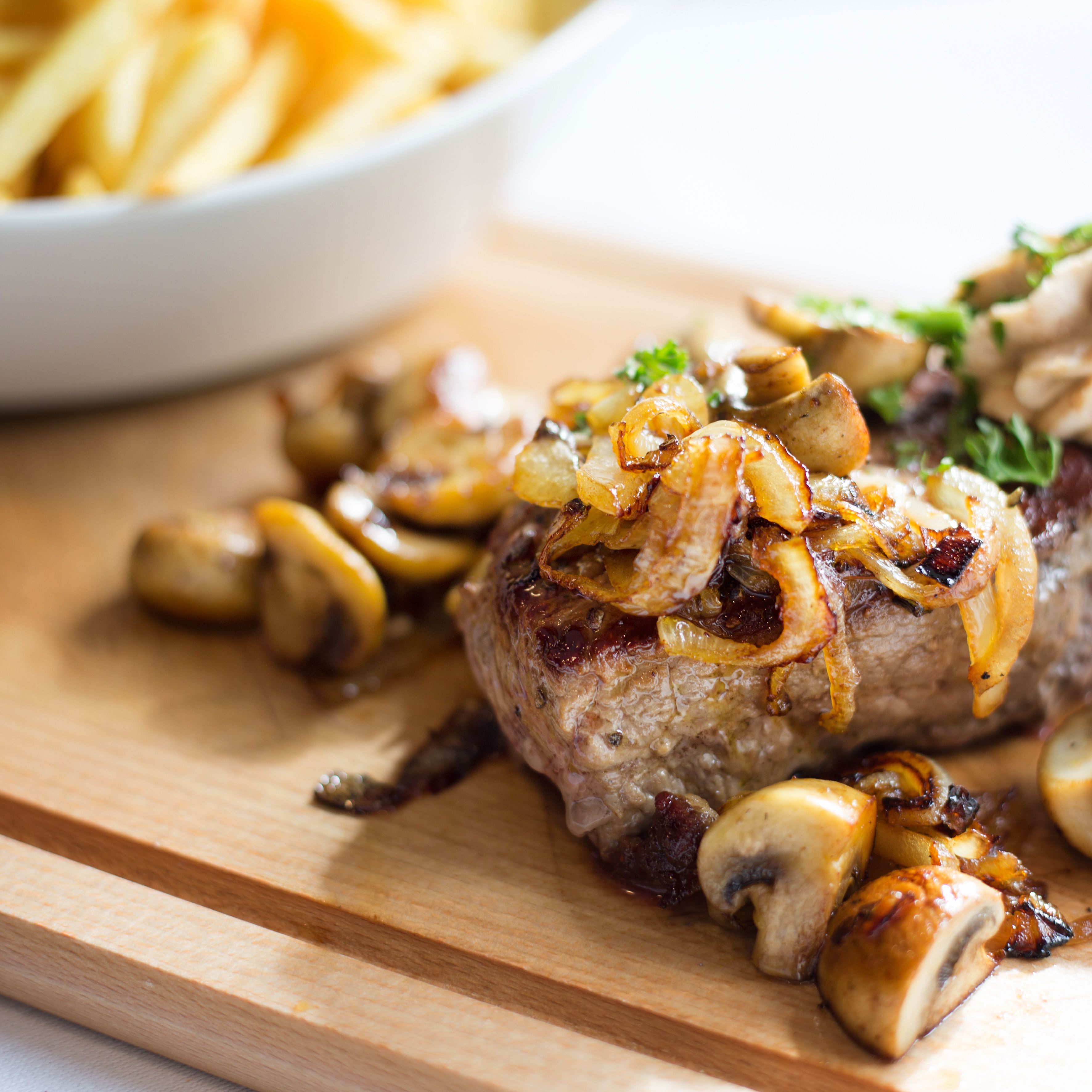 A steak topped with mushrooms and onions, served on a wooden board.