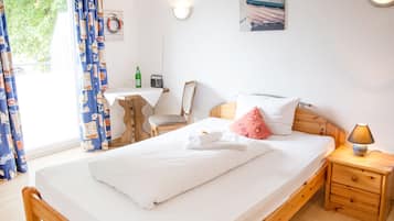 A neatly made bed with white linens, a wooden nightstand with a lamp, a chair, a small table with a radio and a bottle, and a wall-mounted picture.