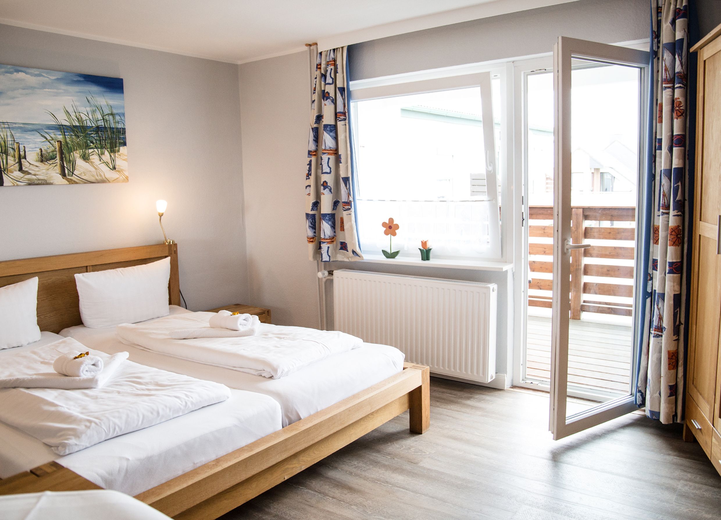 A hotel room with a wooden bed, white linens, a painting of a beach scene, and a view of a wooden deck through an open door.
