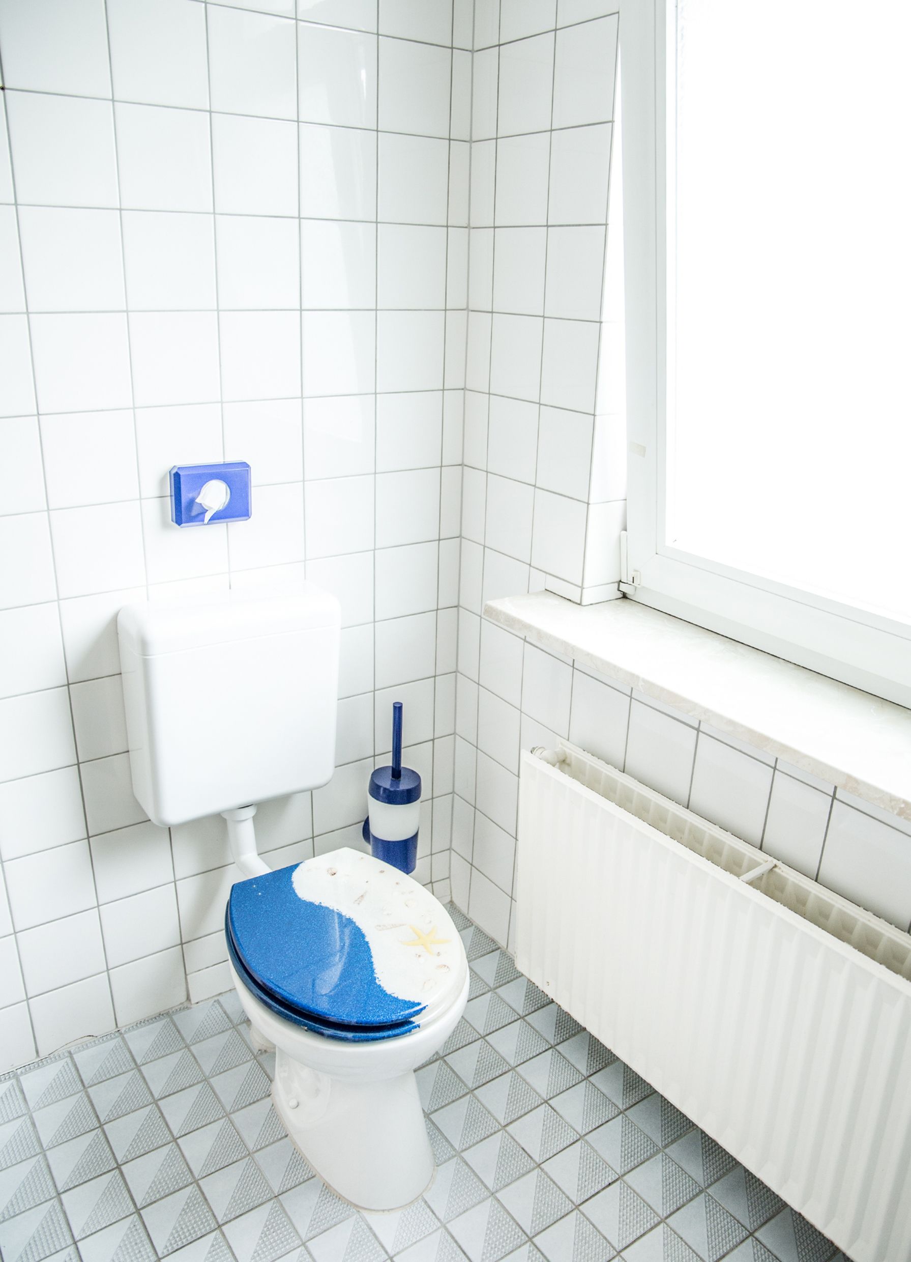 A small, clean bathroom with white tiles, a toilet with a blue seat, a blue toilet brush, and a window with blinds.