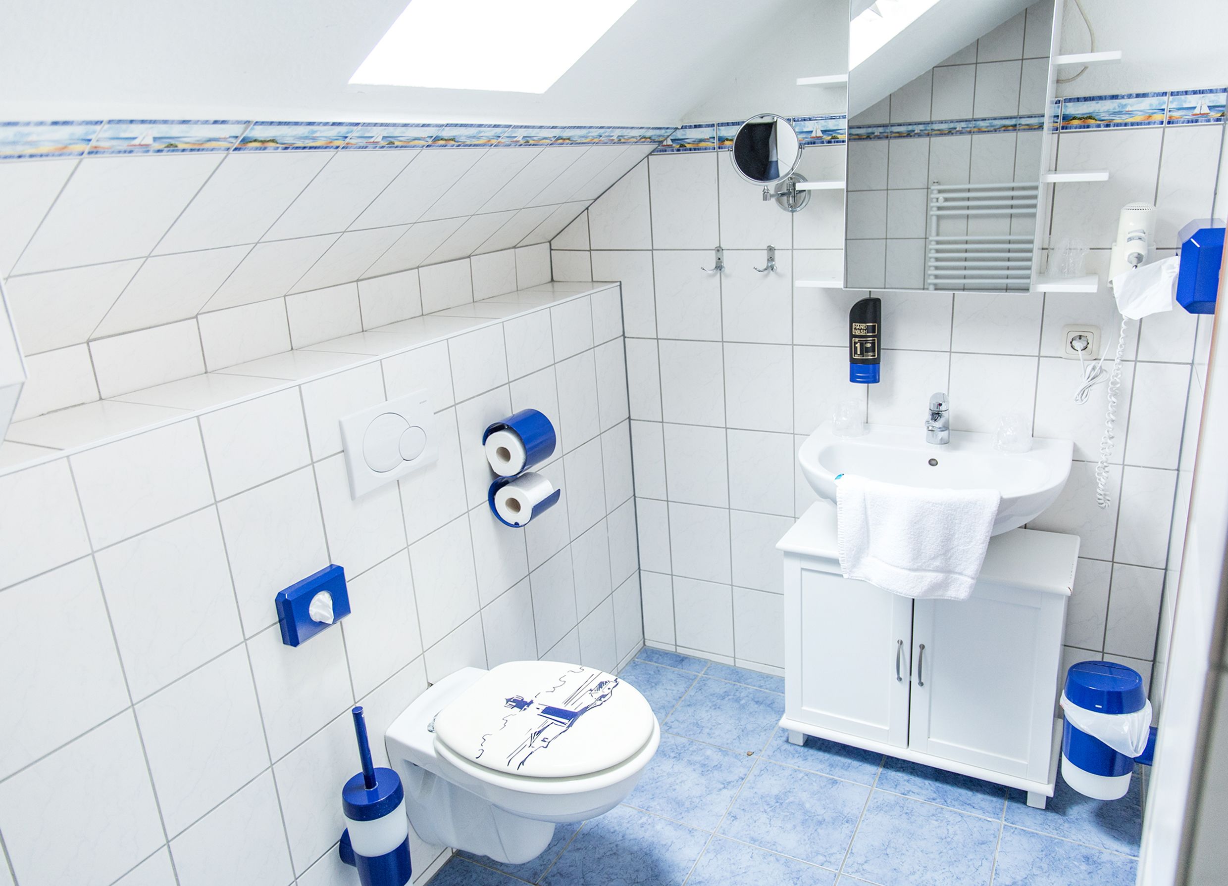 Une petite salle de bain blanche, dotée d’un puits de lumière, de toilettes, d’un lavabo et d’un miroir.