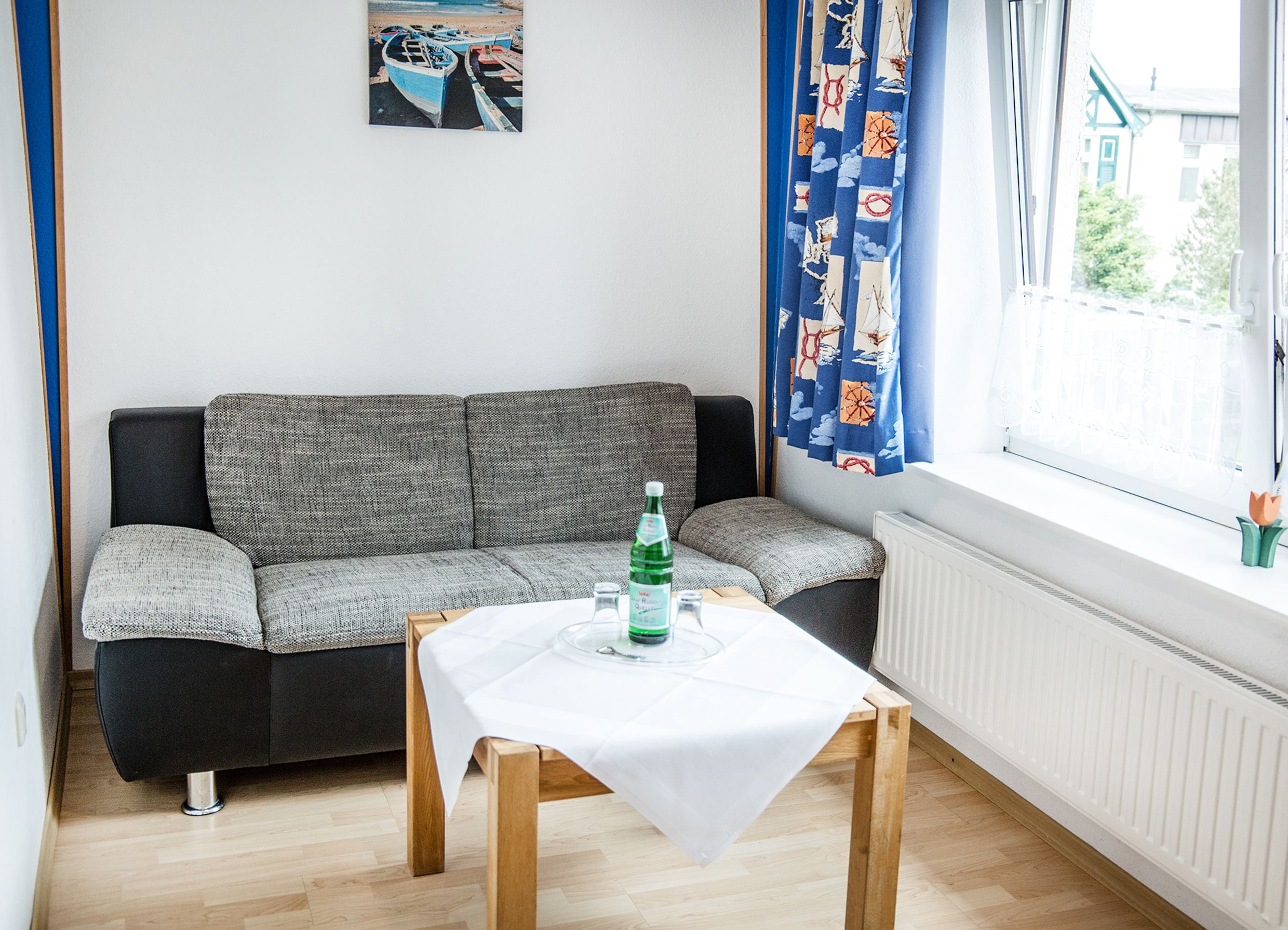 Ein Zimmer mit grauem Sofa, einem kleinen Holztisch mit weißem Tischtuch und einem Fenster mit blauen Vorhängen.